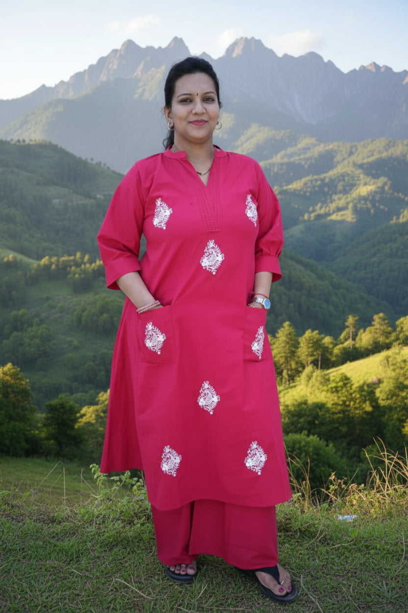 Woman wearing a bright pink traditional outfit with white patterns in a room with light-colored walls and a window.